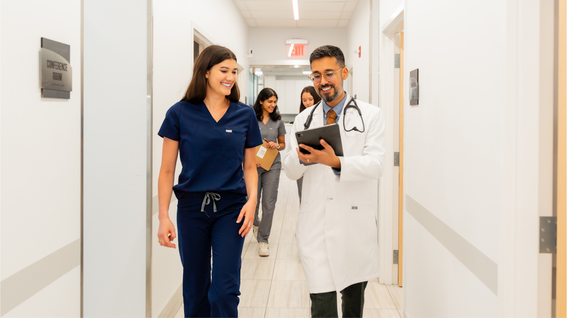 A doctor in a white coat and a nurse in navy scrubs walk together in a bright hallway, smiling and looking at a tablet. Representing Boston Concierge Primary Care, Dr. Jay Luthar leads the team while two staff members follow holding clipboards.
