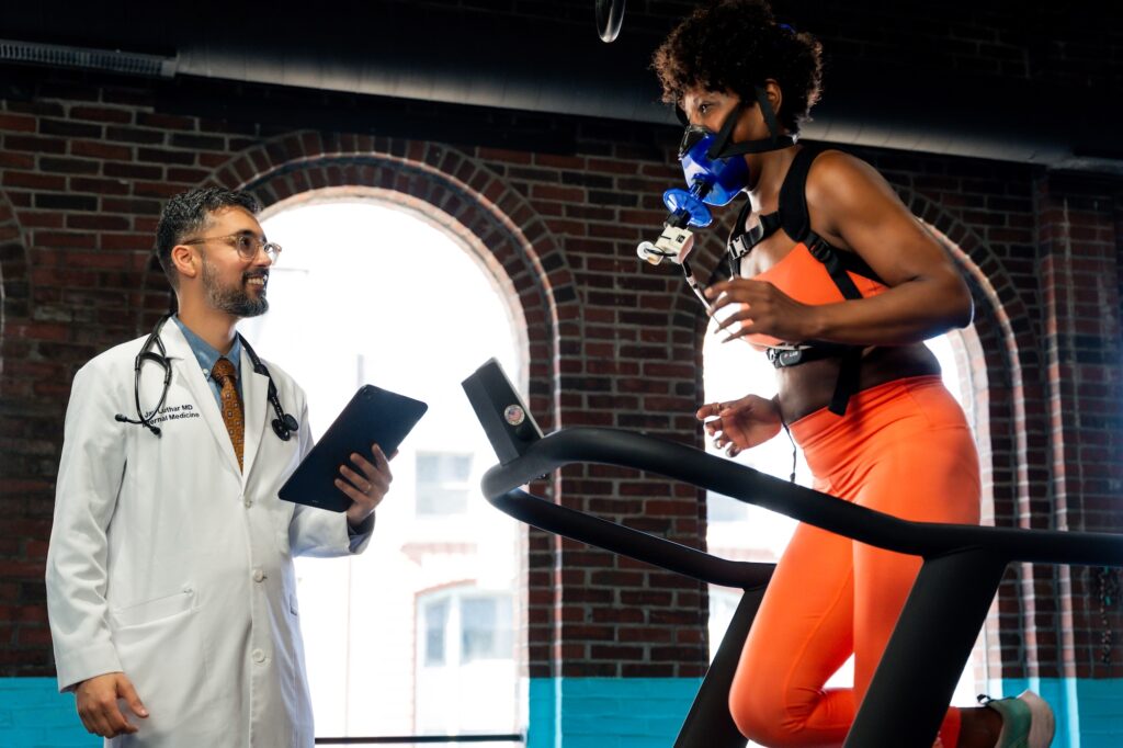 Dr. Julia Loewntha, a Boston Concierge Primary Care Doctor, uses a stethoscope and tablet to observe a woman in orange athletic wear running on a treadmill with a breathing mask, in a bright room with brick walls and large windows.