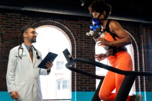 Dr. Julia Loewntha, a Boston Concierge Primary Care Doctor, uses a stethoscope and tablet to observe a woman in orange athletic wear running on a treadmill with a breathing mask, in a bright room with brick walls and large windows.