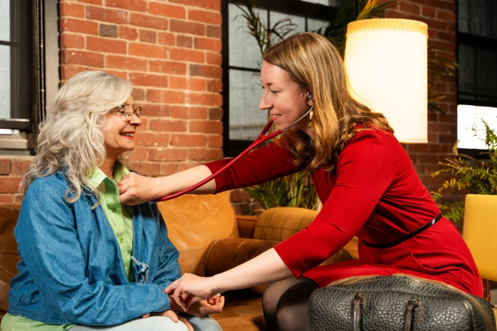 A Boston Concierge Primary Care Doctor in a red dress listens to the chest of an older woman with gray hair and glasses using a stethoscope, both smiling and seated on a cozy couch in a warm brick-walled room.