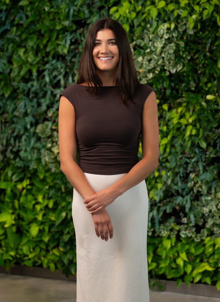 A woman with long dark hair, wearing a sleeveless brown top and a white skirt, stands smiling with her hands clasped in front of a lush green leafy wall.