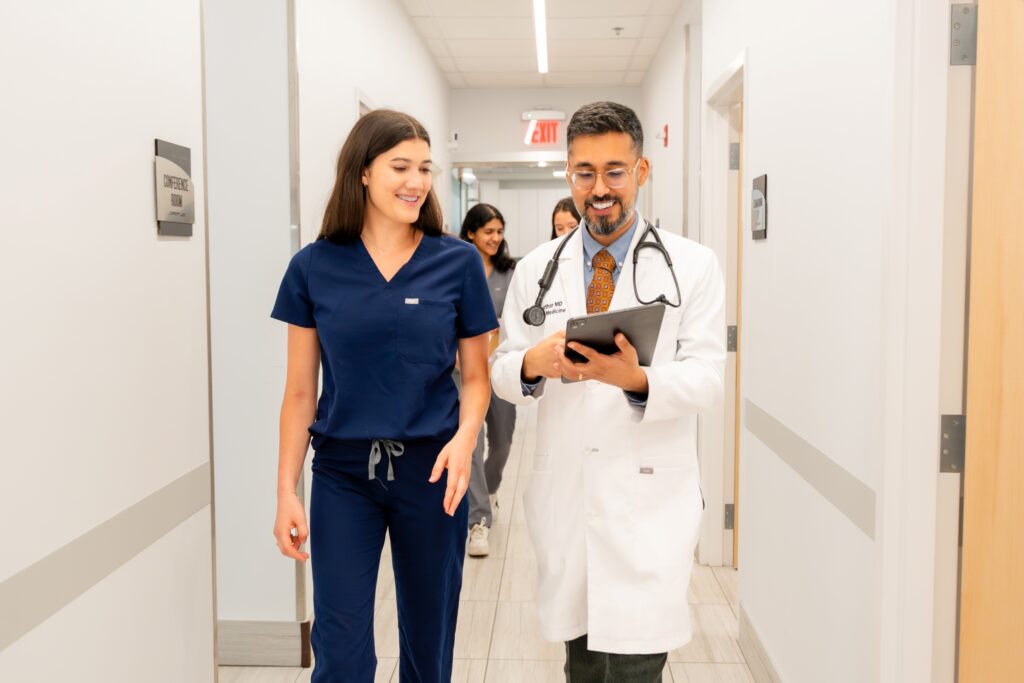 A doctor in a white coat and stethoscope shows a tablet to a smiling nurse in navy scrubs as they walk down a bright hallway; two more people walk behind them.