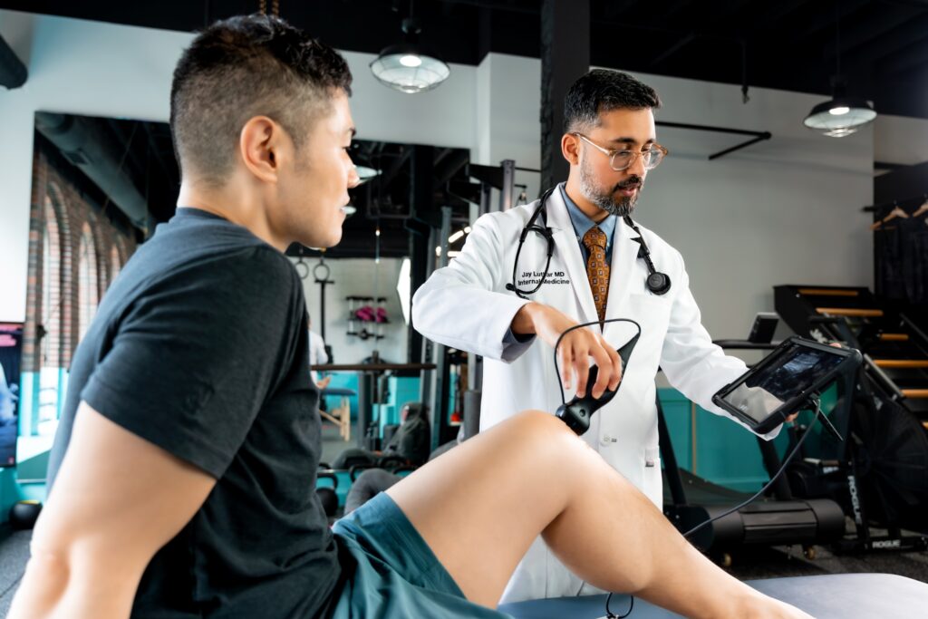 A doctor in a white coat examines a patients knee using a handheld device while viewing results on a tablet in a modern clinic setting. The patient sits on an exam table wearing shorts.