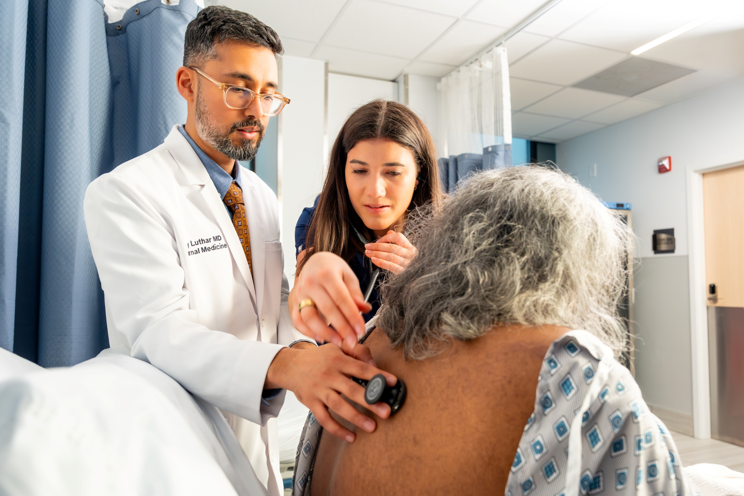 EDITED_LUTHAR_MARCH2025--123 (2) A doctor and a medical student listen to an older patients back with a stethoscope in a hospital room. The patient is wearing a hospital gown and sitting on a bed.