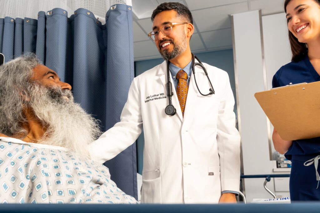 A doctor in a white coat smiles and talks with an older patient in a hospital bed, while a nurse holding a clipboard stands nearby, also smiling. The scene is bright and friendly.