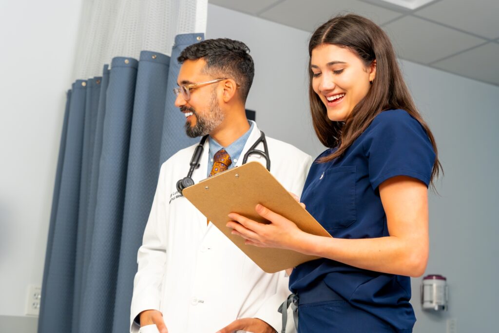 Two healthcare professionals, one in a white coat and the other in navy scrubs, smile and review a clipboard together in a clinical setting with privacy curtains in the background.