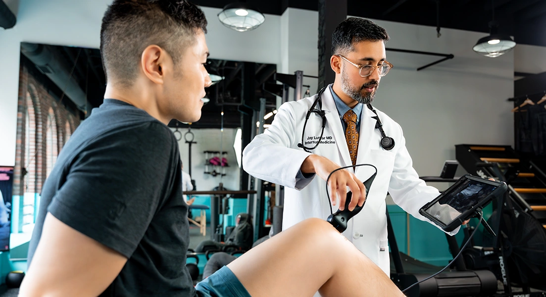 A doctor in a white coat examines a patient’s knee using a handheld device while viewing results on a tablet in a modern medical or physical therapy clinic.