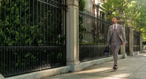 A man in a brown plaid suit carrying a briefcase walks confidently along a sunny, tree-lined sidewalk next to an ornate black iron fence and manicured shrubs.
