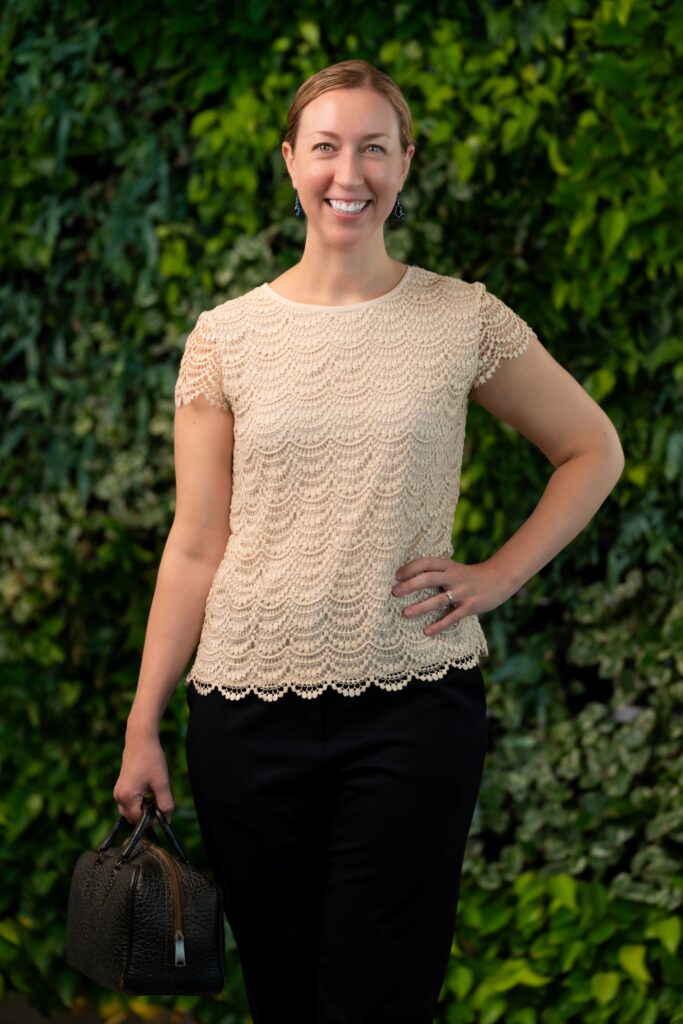 A smiling woman stands in front of a leafy green background, wearing a cream-colored scalloped top and black pants, holding a black handbag in her left hand.