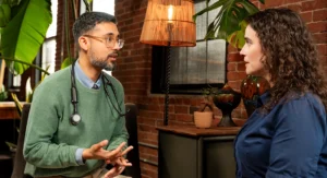 A doctor wearing glasses and a stethoscope talks with a female patient in a cozy office with brick walls, large plants, and warm lighting.