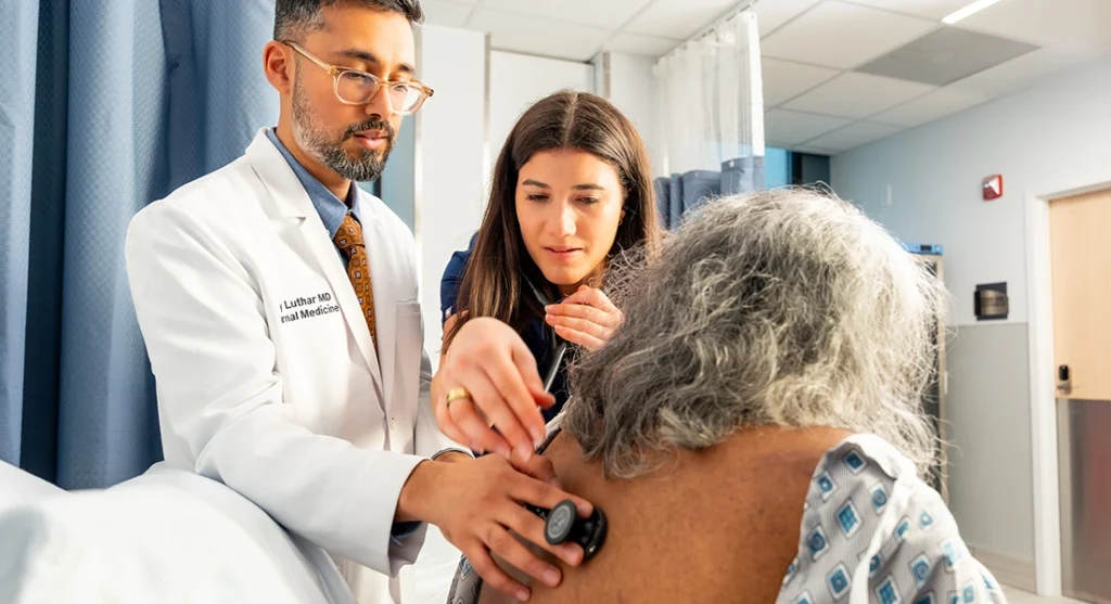 A doctor and a medical student use a stethoscope to examine the back of an elderly patient in a hospital gown, inside a brightly lit medical exam room.