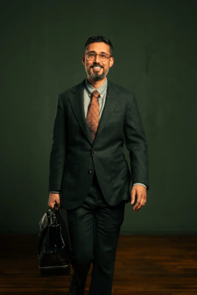 A man wearing glasses and a dark suit with a patterned tie smiles while walking forward, holding a black briefcase in his left hand. The background is plain and dark green.