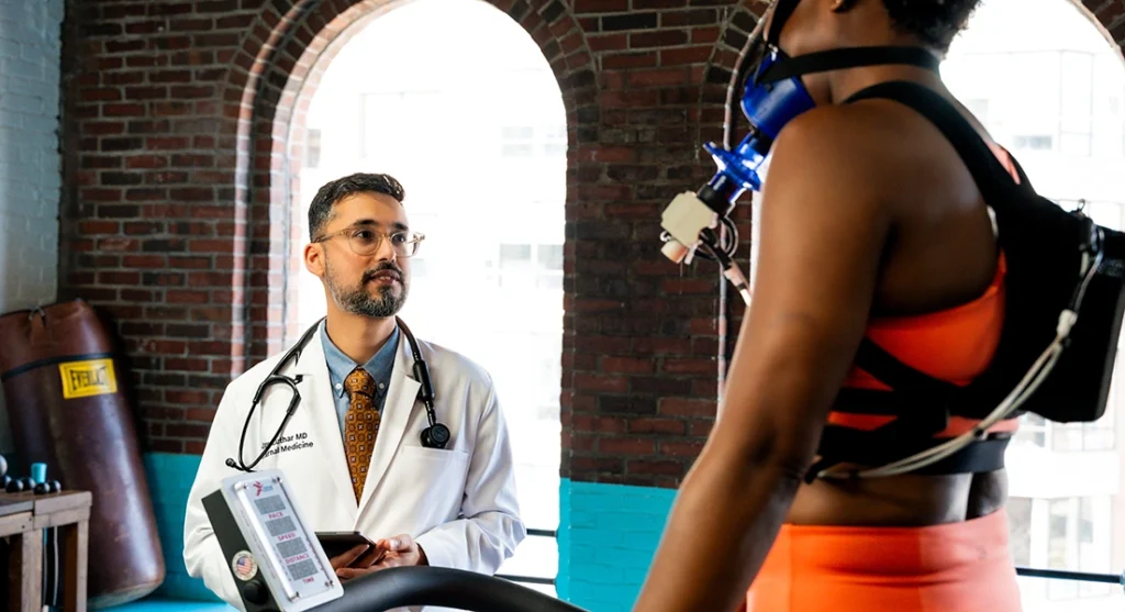 A doctor with a stethoscope observes a woman on a treadmill wearing an orange sports outfit and a breathing mask, monitoring her during an exercise test in a well-lit room with brick walls and large windows.