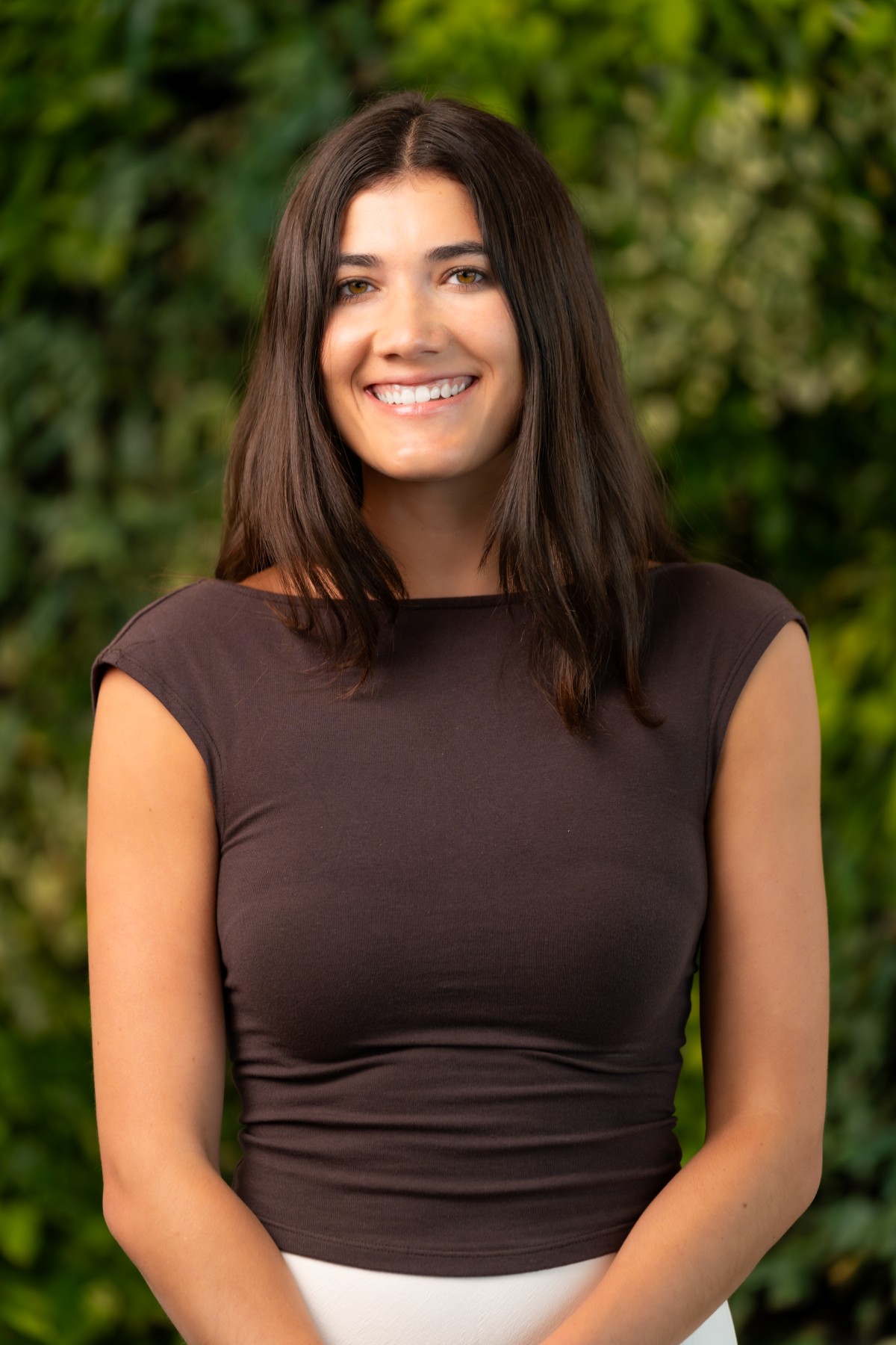 A woman with long dark hair, wearing a short-sleeved dark brown top and white skirt, smiles while standing in front of a leafy green background, representing the friendly concierge medicine team.
