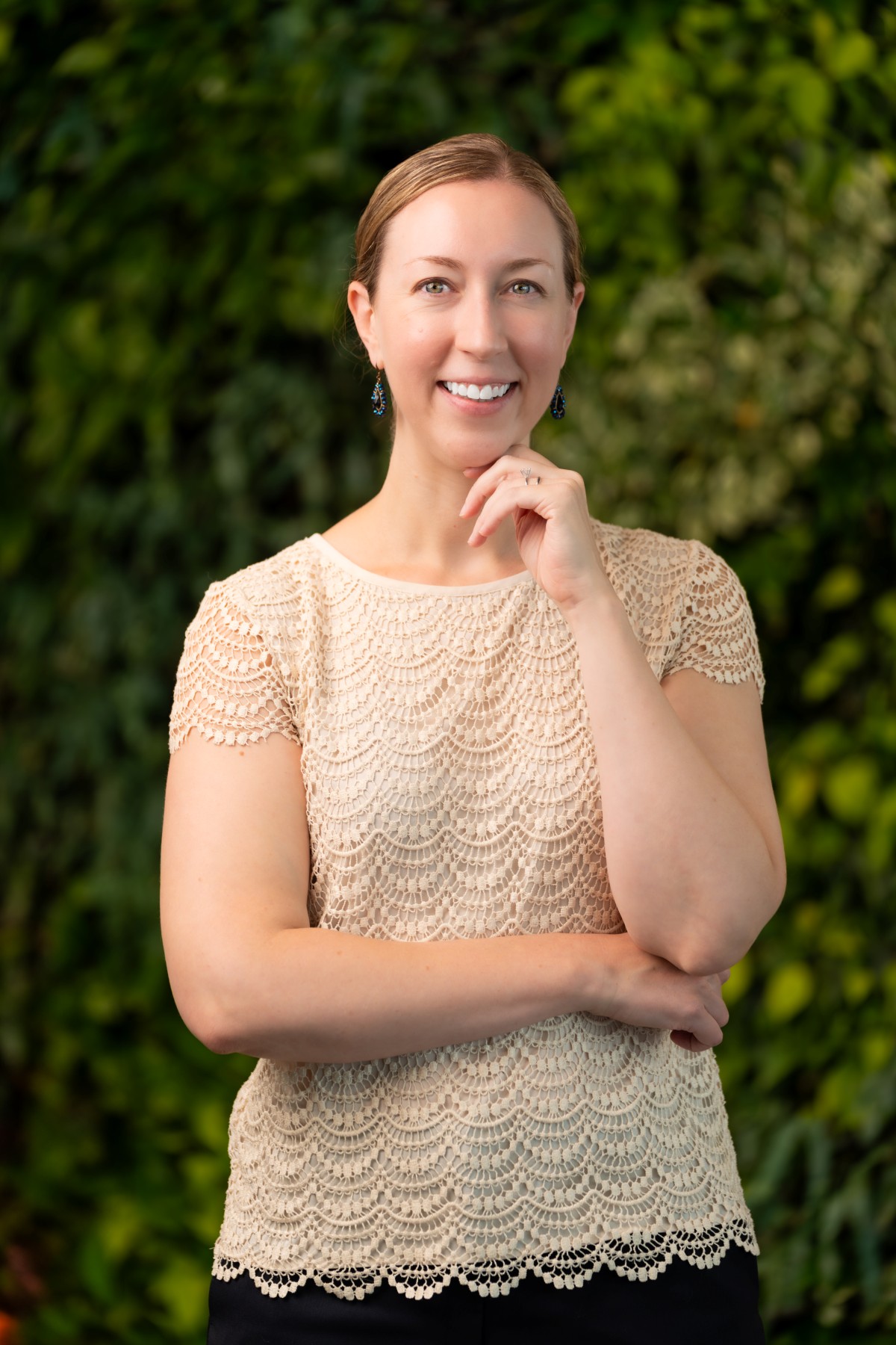 A woman with light brown hair in a low bun is smiling, standing with one arm crossed and the other hand touching her chin. She is wearing a beige lace top and earrings, with a green leafy background like a concierge medicine team portrait.