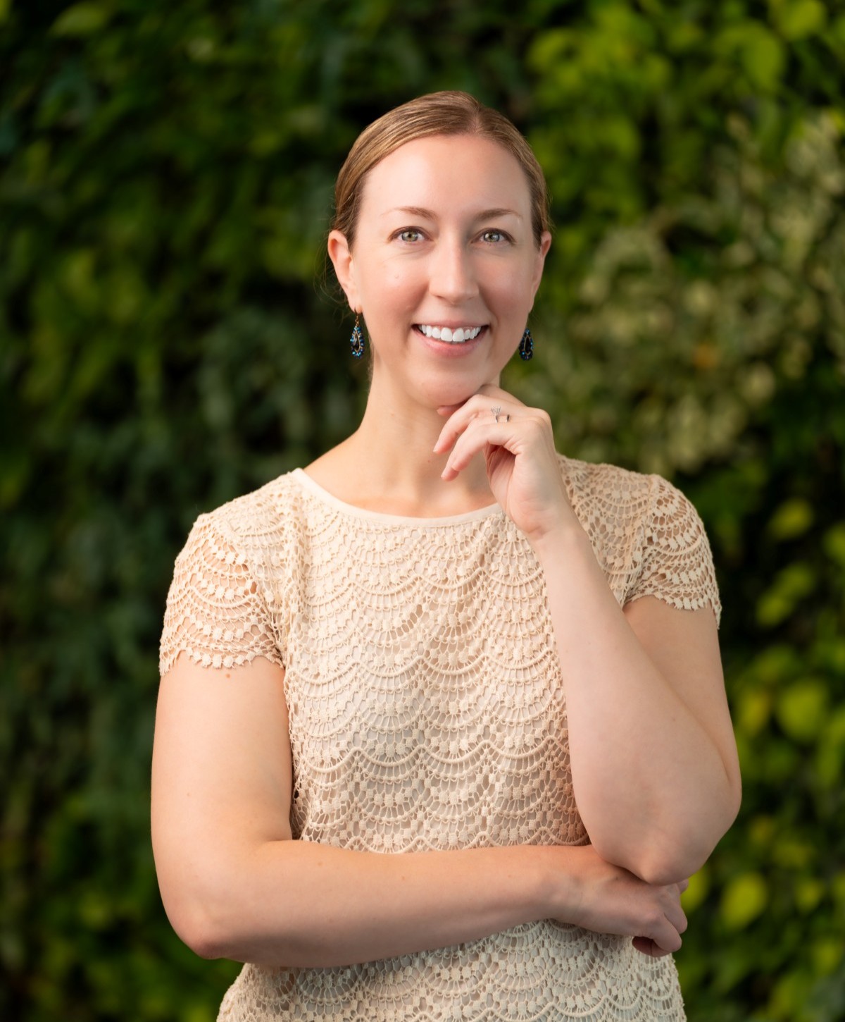 A woman with light brown hair tied back, wearing a cream lace top and blue earrings, stands against a lush green leafy background, smiling with one hand touching her chin and the other arm crossed.