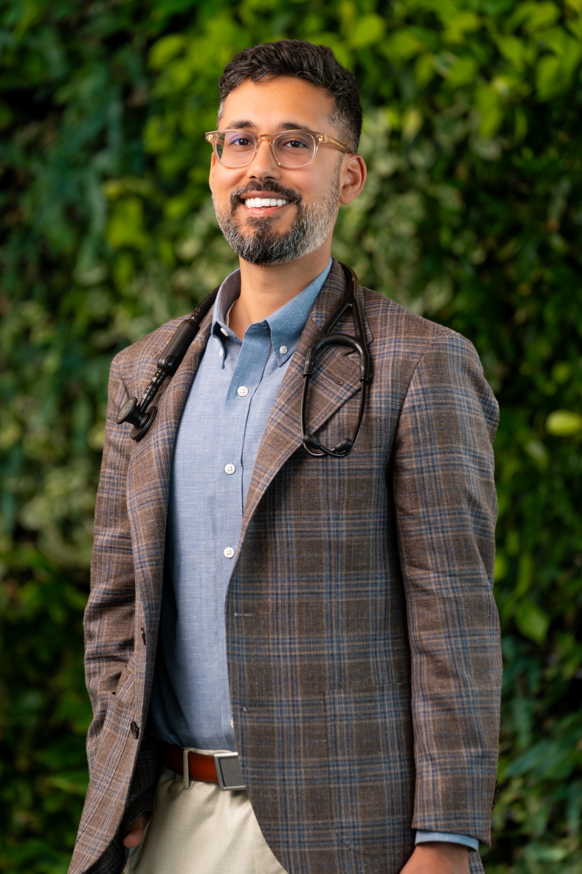 A smiling man with glasses and a beard, part of a concierge medicine team, wears a plaid blazer, blue shirt, and stethoscope draped around his neck, standing in front of a leafy green background.