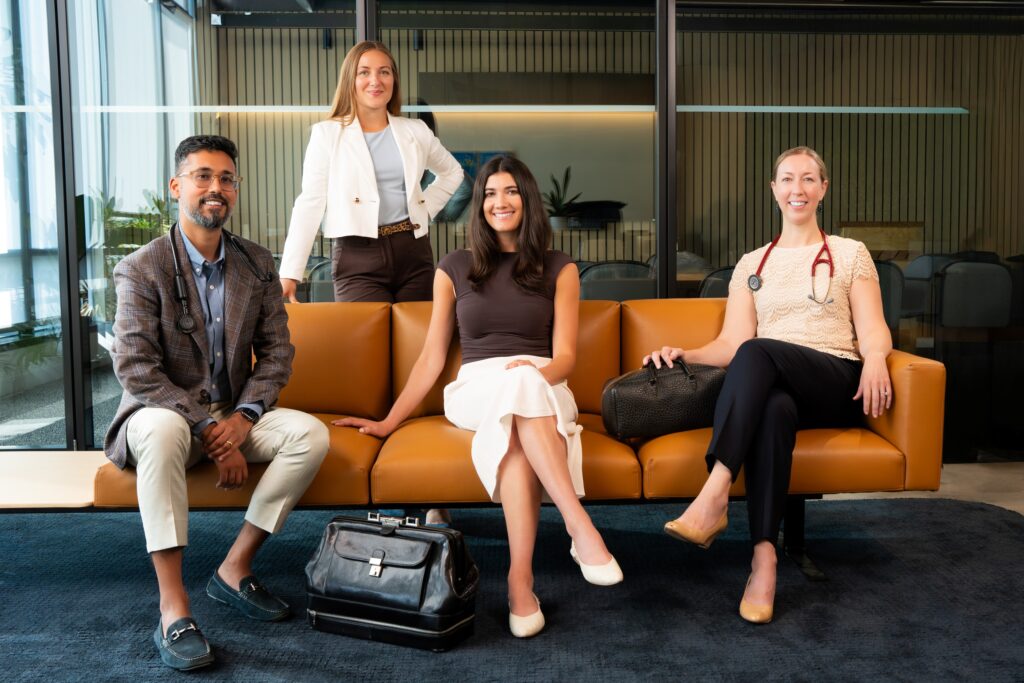 Four medical professionals, three women and one man, sit and stand around a modern tan sofa in an office. Two have stethoscopes, and all are smiling—an approachable concierge medicine team in business or business-casual attire.