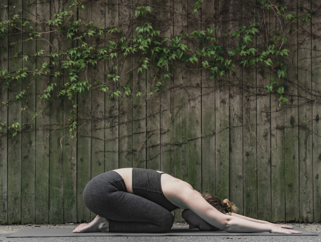 A person in dark athletic wear practices child’s pose on a yoga mat outdoors, in front of a wooden fence with green vines, embracing mindful movement as recommended by Dr. Julia Loewenthal, MD.