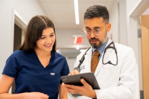 A doctor in a white coat and stethoscope shows a digital tablet to a smiling nurse in navy scrubs as they stand together in a brightly lit hospital hallway.