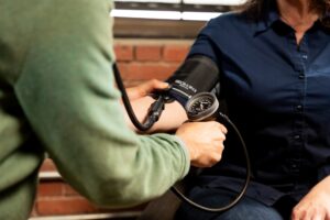 A person in a green sweater measures another’s blood pressure with a manual cuff and gauge, while a HEPA air purifier runs nearby—an added step for supporting cardiovascular health. The person being tested wears a navy blue shirt.