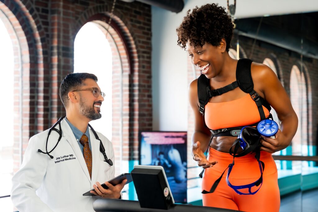 A doctor in a white coat smiles while discussing VO₂ Max Testing with a woman in orange workout clothes holding a water bottle and weights, standing in a bright gym with large windows in the background.