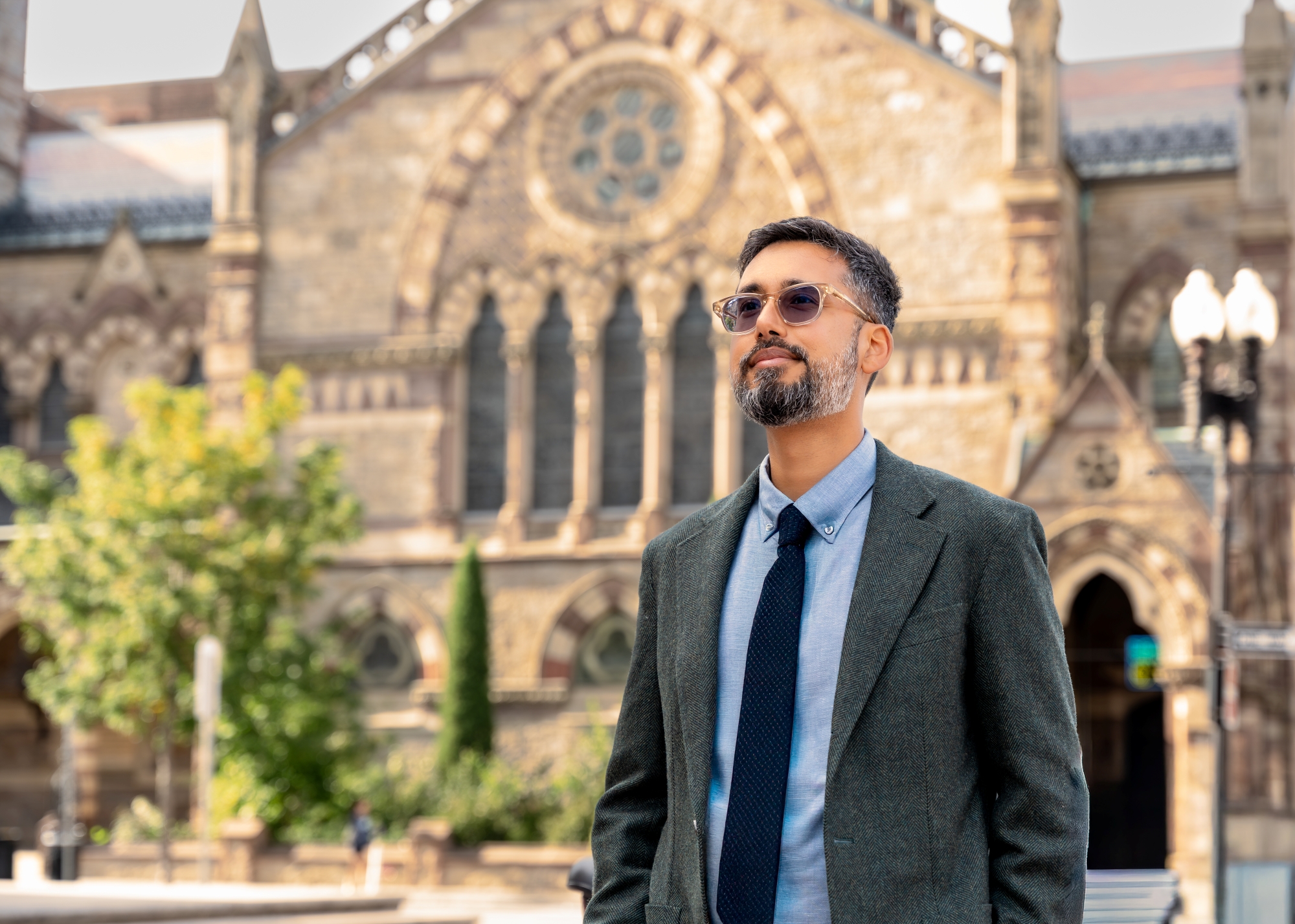 Dr. Luthar wearing sunglasses, a suit, and a tie stands outdoors, smiling in front of a historic stone building with arched windows and detailed architecture, radiating confidence and vitality that reflect his commitment to longevity health.