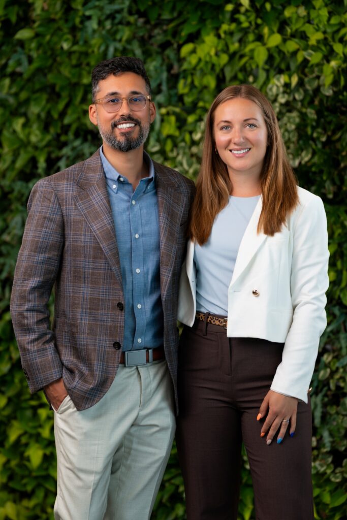 A man and a woman stand side by side, smiling at the camera. The man wears glasses, a blue shirt, and a plaid blazer. The woman wears a white blazer over a light blue top. Green foliage is in the background.