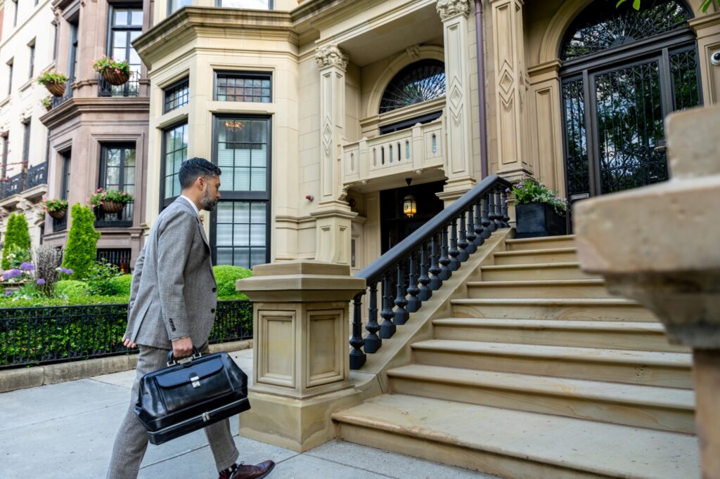 A man in a suit carrying a black briefcase walks up the steps to an elegant, historic building with large windows and ornate architectural details.