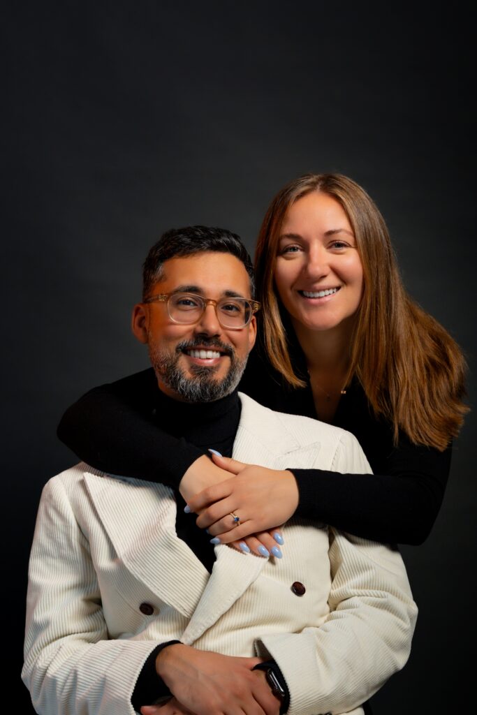 A smiling woman with long brown hair hugs a bearded man in glasses and a white jacket from behind. Both are happy, posing together against a dark background. The woman rests her hand on the mans shoulder.
