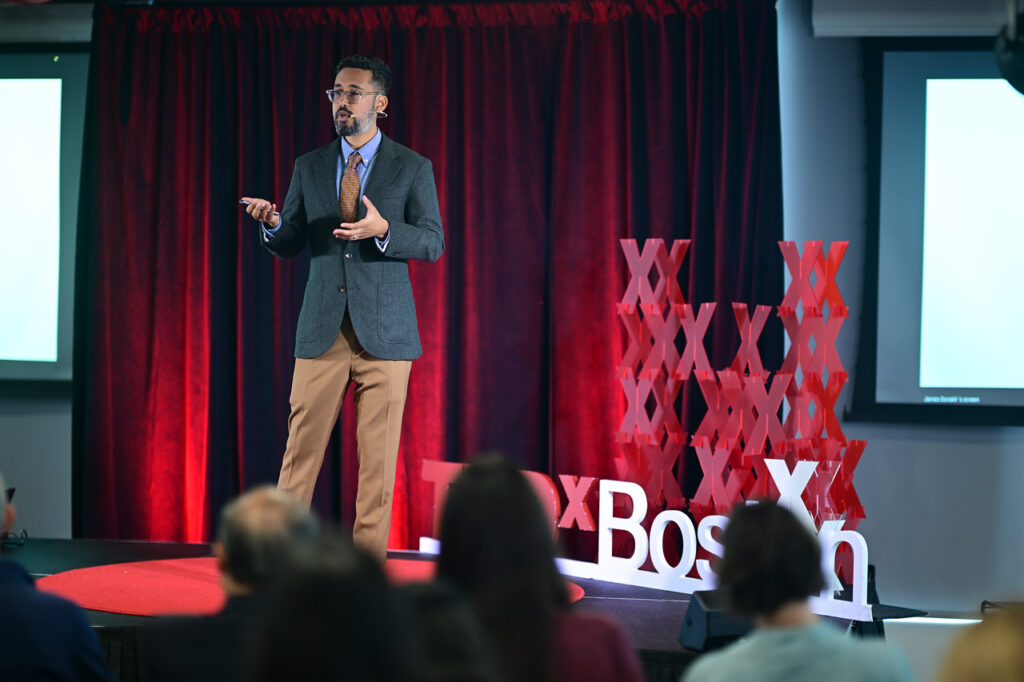 A man in a suit and tie speaks on stage at a TEDxBoston event, gesturing with one hand. The stage features red TEDx letters and decorative X shapes, with an audience visible in the foreground.