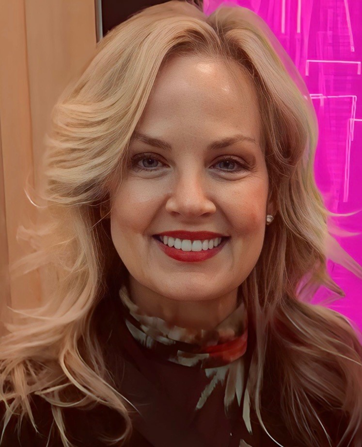 CLTPHOTO A woman with long, wavy blonde hair and a bright smile stands in front of a neon pink background, wearing a dark floral-patterned top.