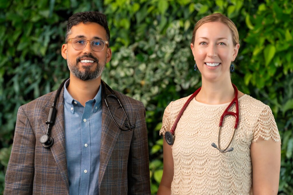 Dr. Jay Luthar and Dr. Julia Loewenthal, both wearing stethoscopes, standing side by side in front of a green foliage wall.