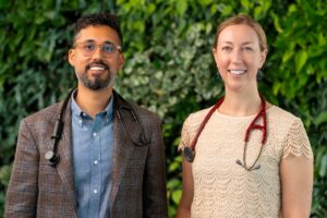 Dr. Jay Luthar and Dr. Julia Loewenthal, both wearing stethoscopes, standing side by side in front of a green foliage wall.