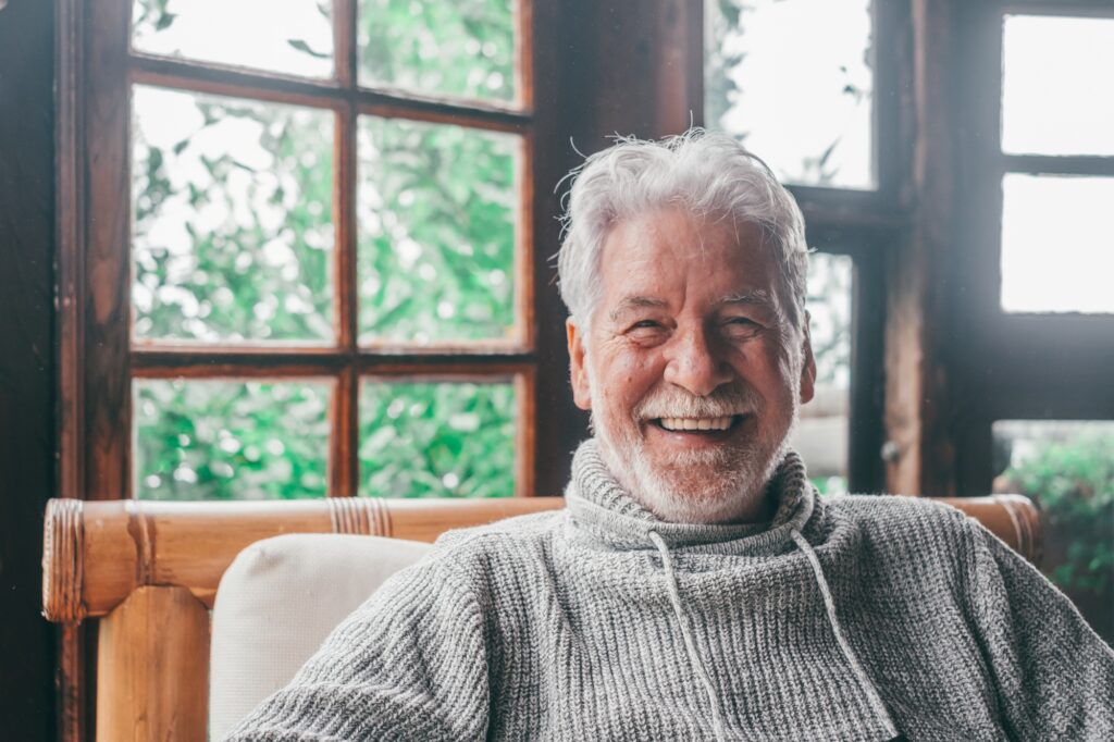 An older man with white hair and a beard, known for advocating lithium’s potential in Alzheimer’s care, smiles warmly while sitting indoors on a wooden chair. He is wearing a gray sweater, with large windows and green foliage visible outside.
