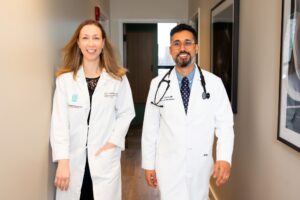Dr. Jay Luthar and Julia Loewenthal MD in white lab coats, one woman and one man, walk down a hallway smiling. The man wears a stethoscope around his neck. They appear confident and professional in a medical office setting.