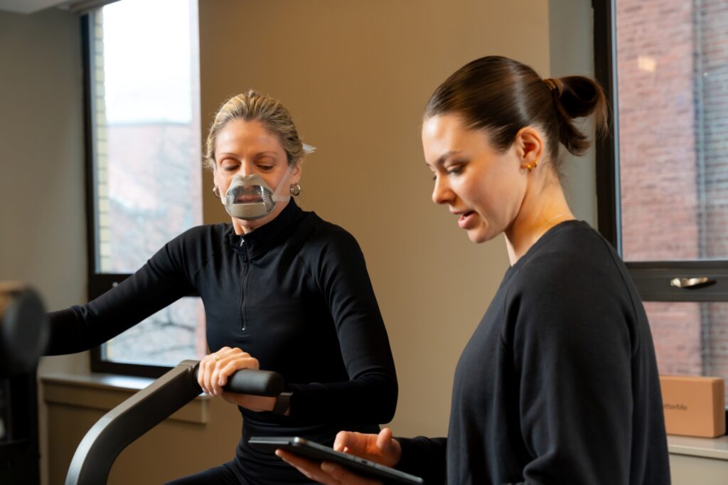 Izzy Antonelli reviews a tablet while a masked patient exercises on a stationary bike in a gym setting.