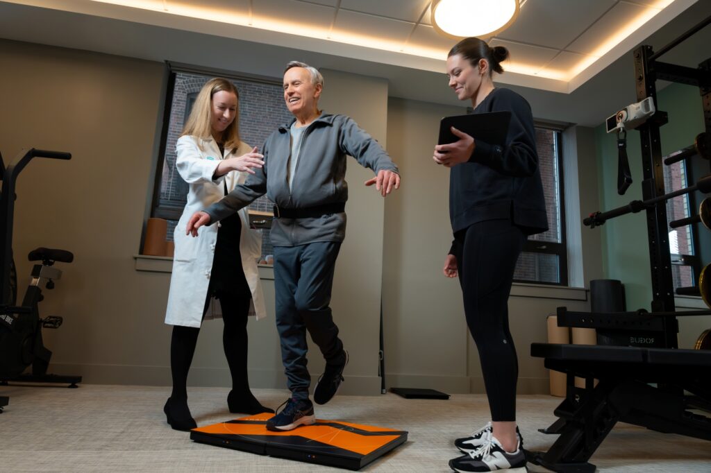 An older man balances on one foot on an exercise platform while a woman in a lab coat assists him and another woman holds a tablet, observing in a gym or physical therapy room.