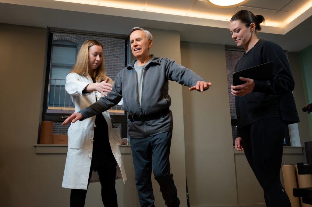 An older man practices balancing on one leg with help from Julia Loewenthal MD in a white coat, while Izzy Antonelli observes and takes notes on a tablet in a clinical setting.