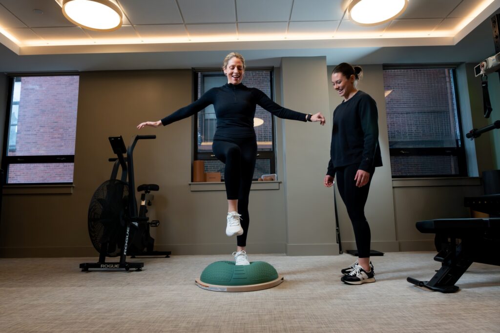 Two women in athletic wear are in a gym. One woman is balancing on one leg on a green half-ball balance trainer, arms outstretched and smiling, while Izzy Antonelli stands nearby, watching and smiling.
