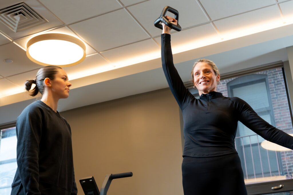 A woman in black athletic wear lifts a kettlebell overhead while smiling, as Izzy Antonelli in similar attire stands nearby watching, inside a brightly lit gym with large windows.