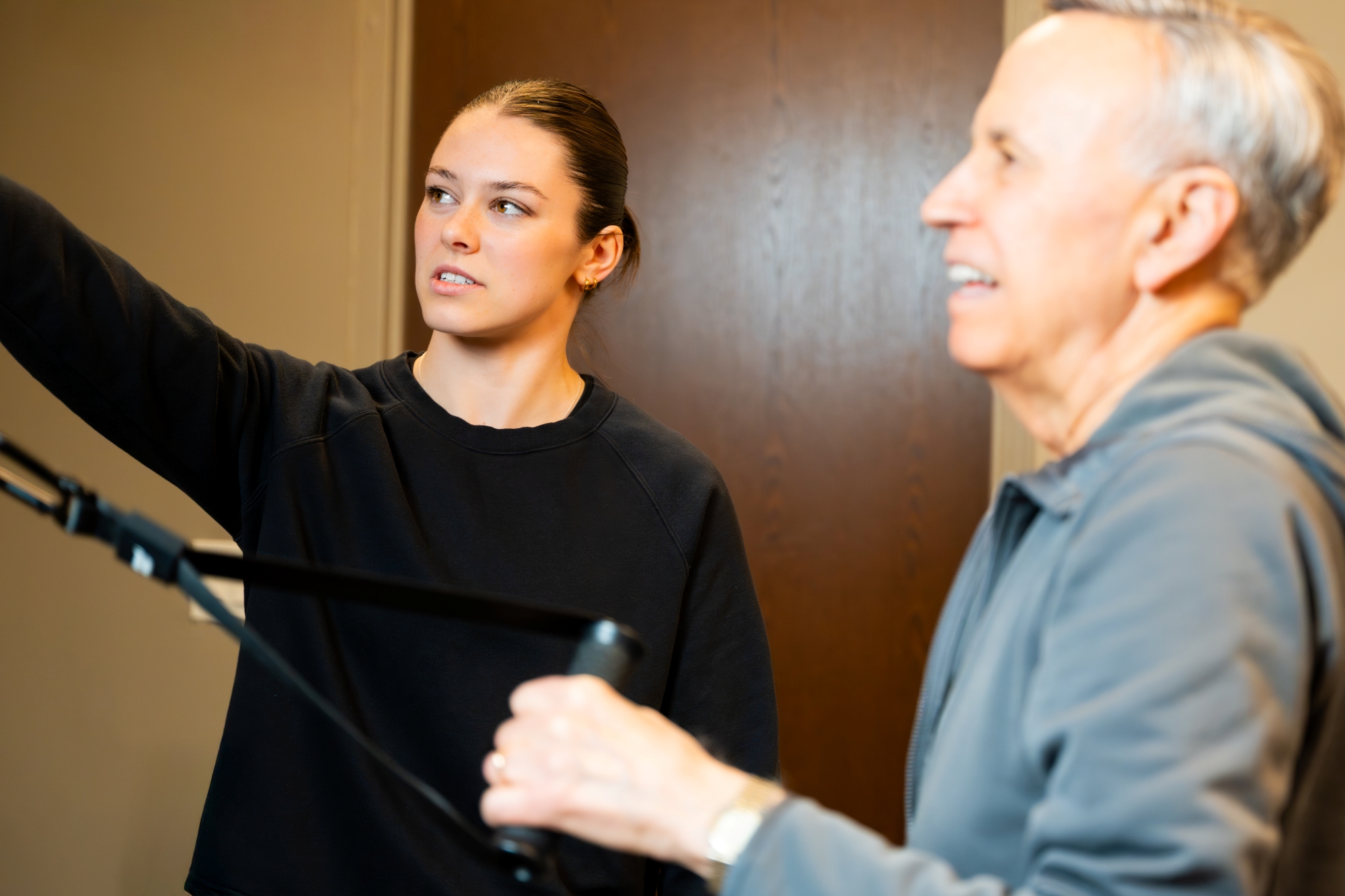 LUTANEN_FEB_2026-Gym_0861-Edit-Gym- Izzy Antonelli instructs an older man using exercise equipment. The woman points ahead while the man holds resistance bands, both focused, in an indoor setting.
