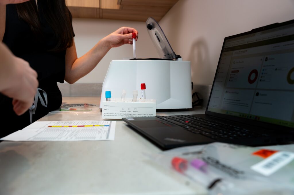 A person uses a lab instrument while another stands nearby; test tubes, paperwork, and a laptop displaying data are on the counter, suggesting a scientific or medical testing environment.