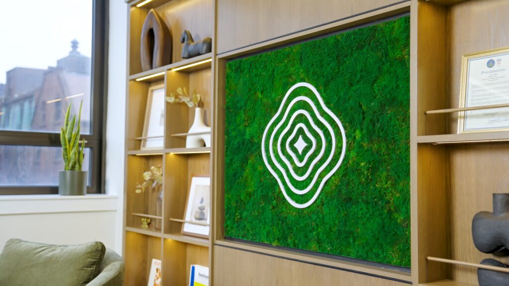 A modern office features a green wall panel with a white abstract line design, surrounded by wooden shelves displaying decor, books, framed photos, and a plant near a window.