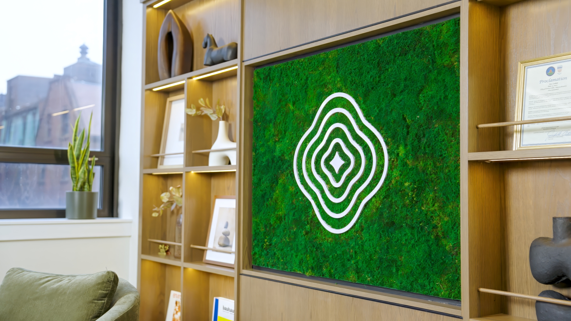 Lutanen features a green wall panel with a white abstract line design, surrounded by wooden shelves displaying decor, books, framed photos, and a plant near a window.