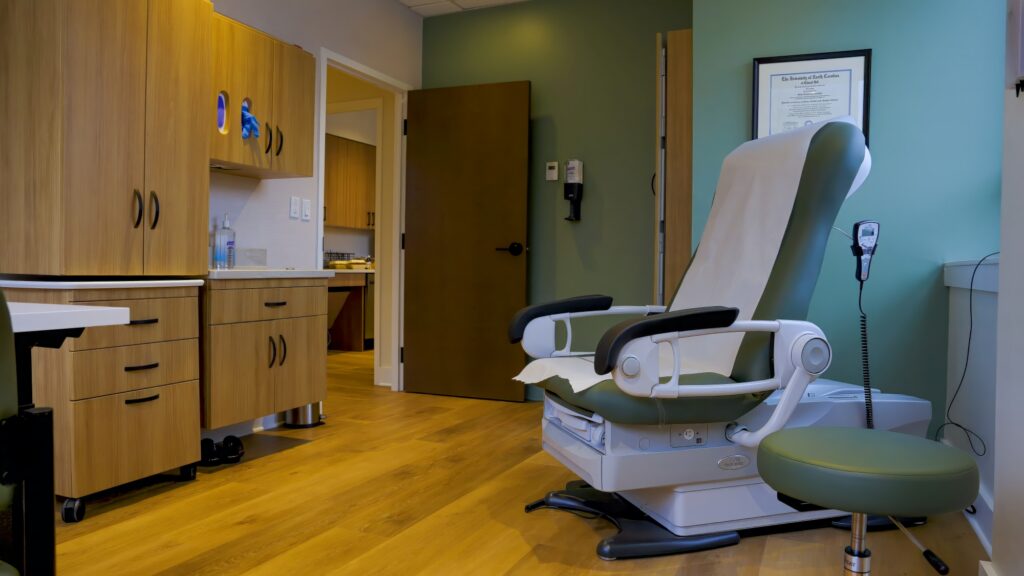 A modern medical exam room with a reclining examination chair, a green stool, wood cabinets, and a framed certificate on a green wall. The room has wood flooring and an open door leading to another area.