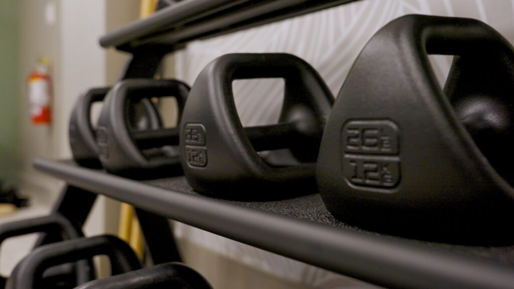 Close-up of black kettlebells arranged on a rack, with visible labels showing weight in pounds and kilograms. The background includes a wall with a fire extinguisher partially visible.