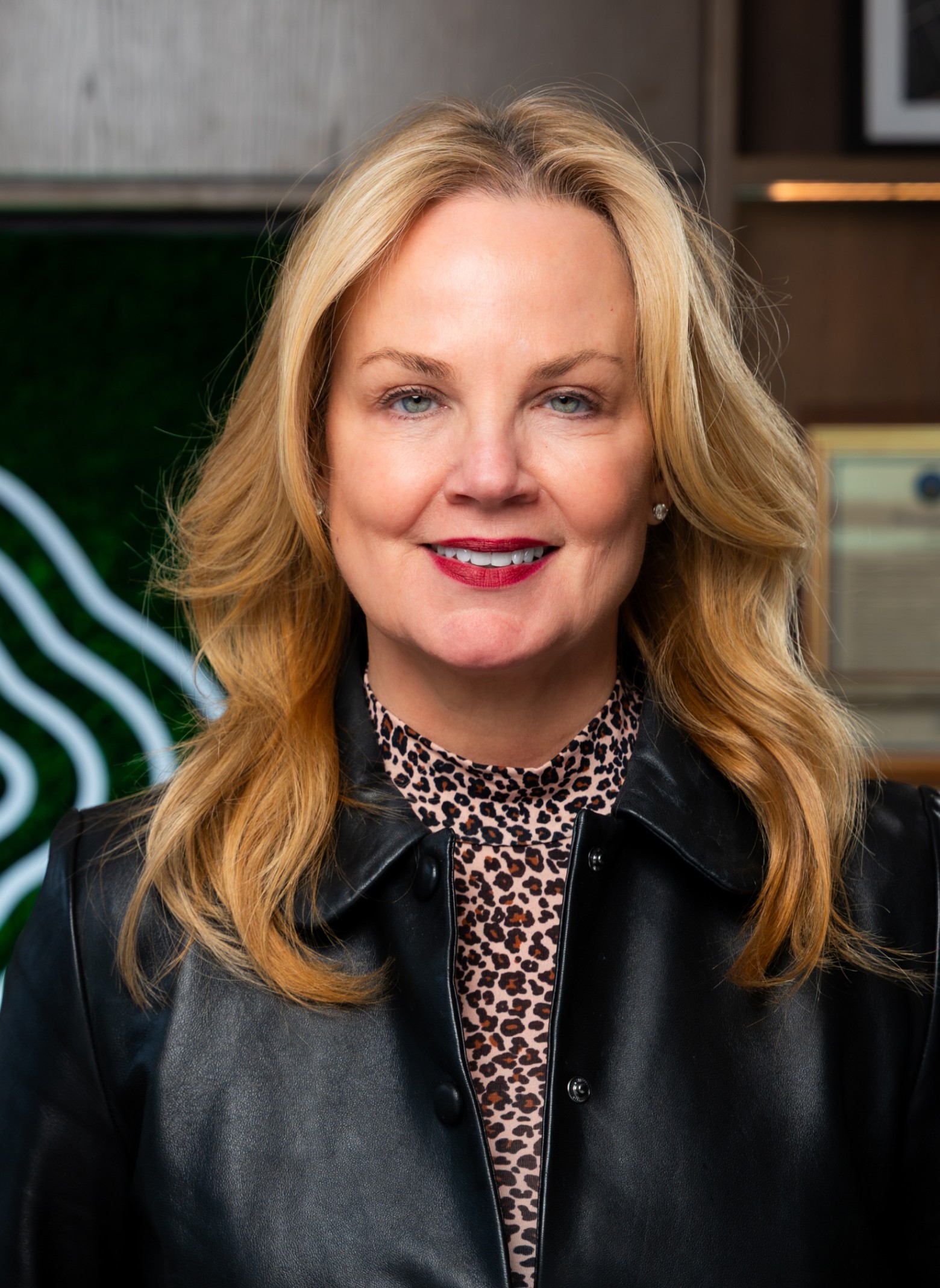 image_Charlene A woman with long blonde hair is smiling at the camera. She is wearing a black leather jacket over a leopard print top. The background features a green and wood panel wall with framed items.
