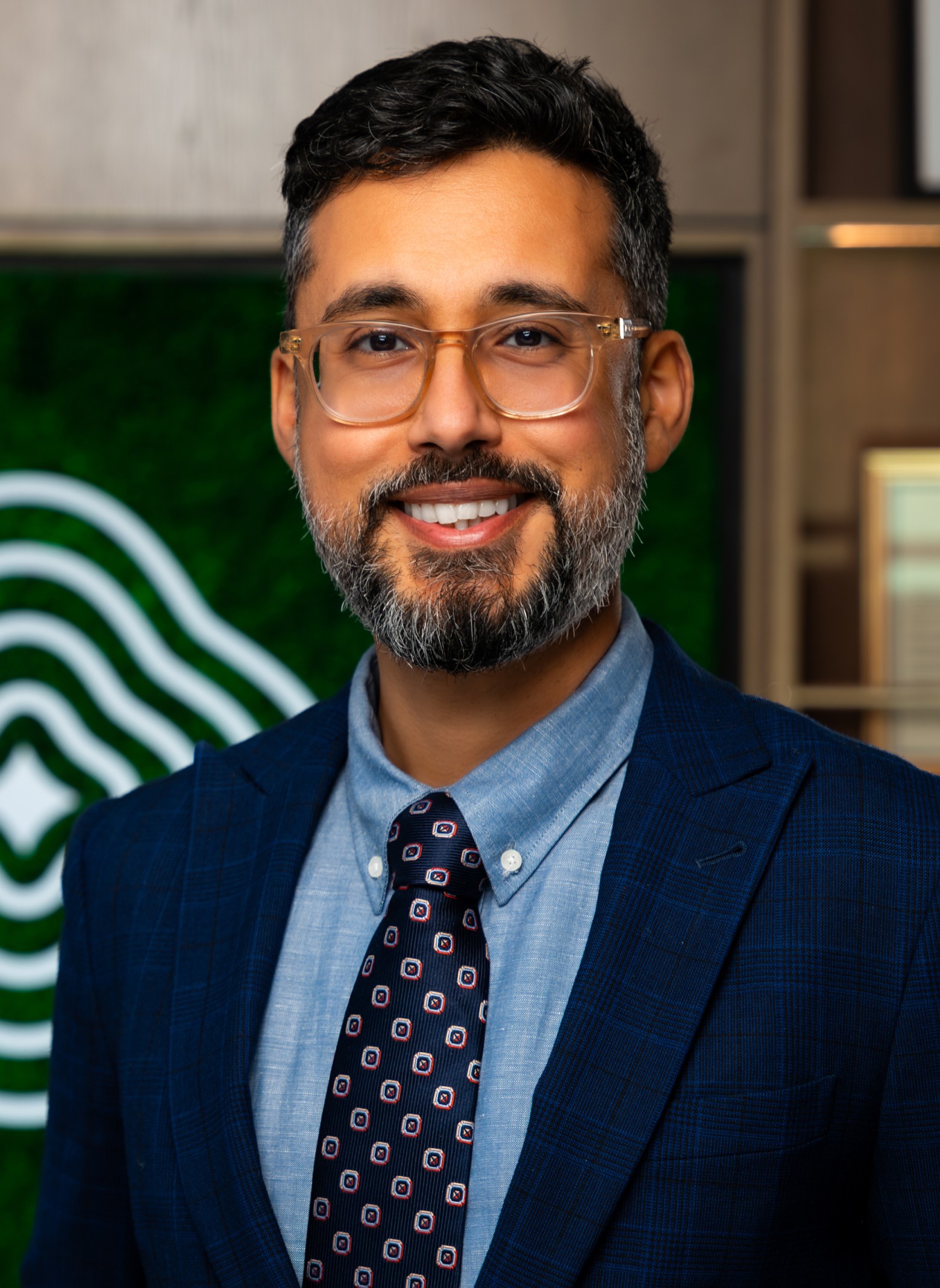 image_Jay Dr. Jay Luthar dark hair, a beard, and glasses smiles while wearing a blue plaid suit, blue shirt, and patterned tie, standing indoors with a green and white abstract logo in the background.