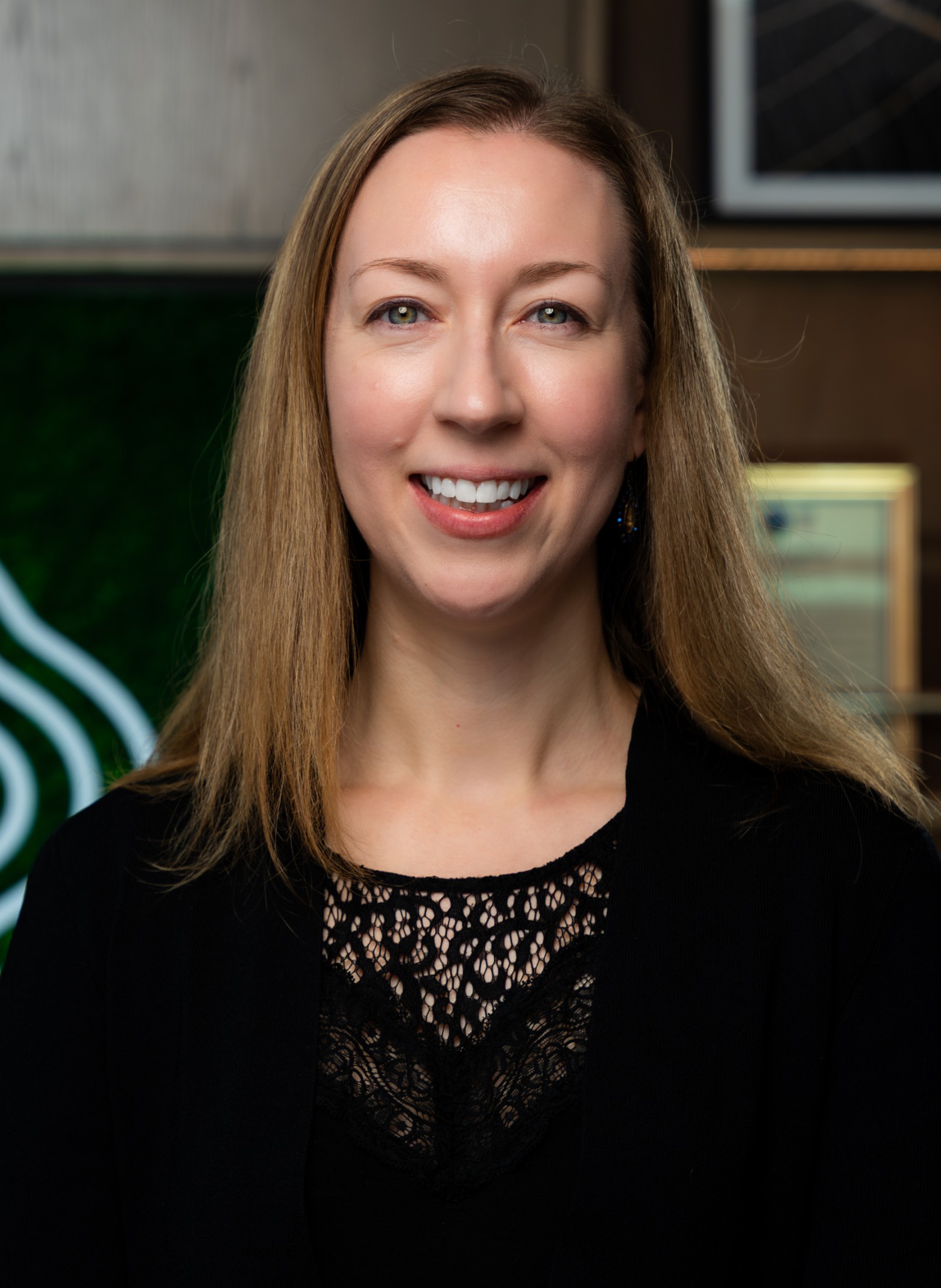 image_Julia A woman with long light brown hair, wearing a black lace top and black cardigan, smiles at the camera. The background features green and brown tones with framed objects on the wall.